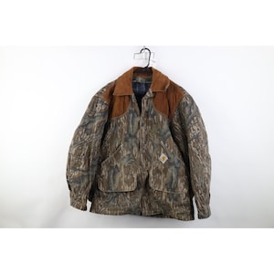 Puede incluir: Una chaqueta Carhartt con cuello de pana marrón y un patrón de camuflaje en tonos marrones, verdes y grises. La chaqueta incluye dos grandes bolsillos delanteros con solapa y un pequeño parche con el logo de Carhartt en el bolsillo inferior.