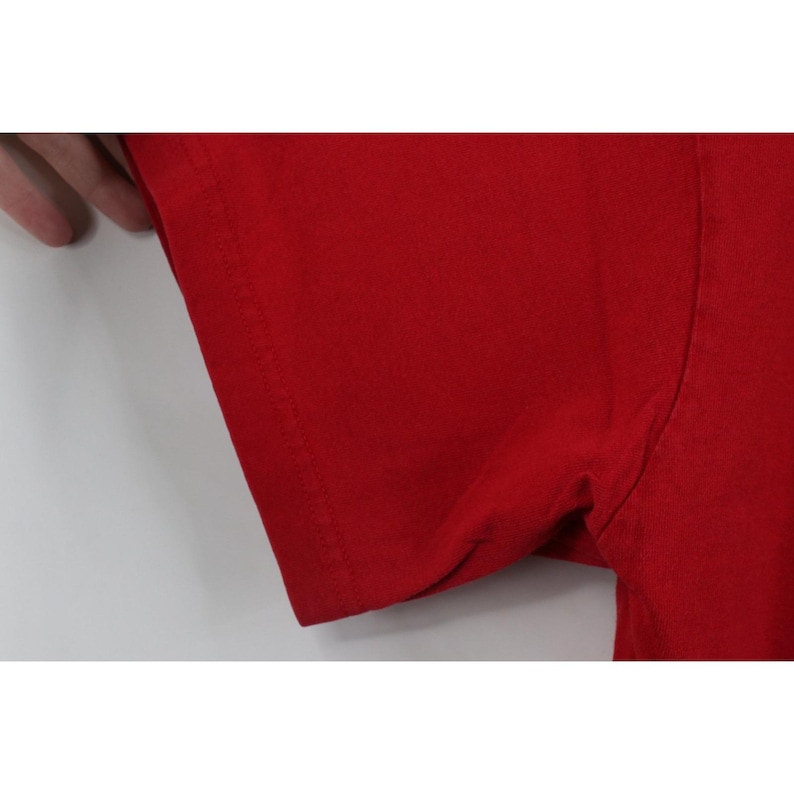 May include: Close-up of a red t-shirt sleeve. The fabric is a solid, vibrant red, and the sleeve's edge is neatly hemmed with a visible seam. The image focuses on the texture and color of the garment.