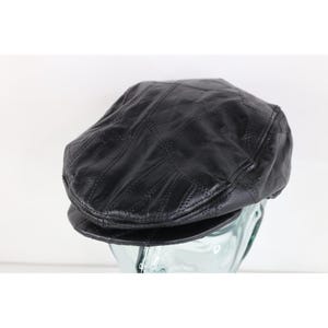 May include: A black leather flat cap constructed from multiple stitched panels. The hat features a smooth, slightly textured surface with visible seams connecting the individual leather pieces, a rounded crown, and a short, stiffened front brim.