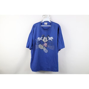 May include: Blue t-shirt featuring a graphic of Mickey Mouse with the text "Walt Disney World". The design includes Mickey's face and arms, with the words "Walt Disney World" in a stylized font. The shirt is hanging on a white hanger.