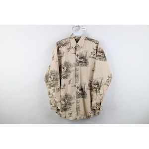 May include: A long-sleeve button-down shirt in a light tan colour featuring an all-over dark brown print of deer, trees, and forest landscapes. The shirt includes a standard collar, a single chest pocket, and buttoned cuffs on the sleeves.