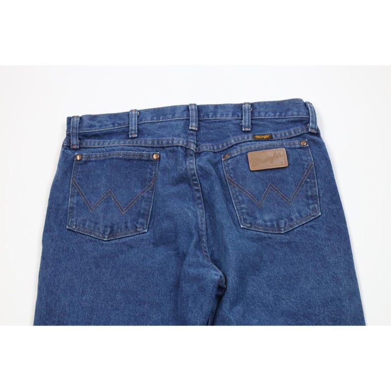 May include: Close-up of blue denim jeans with the Wrangler logo on the back pocket. The jeans have a classic five-pocket design with a straight leg cut. The denim is a medium wash.