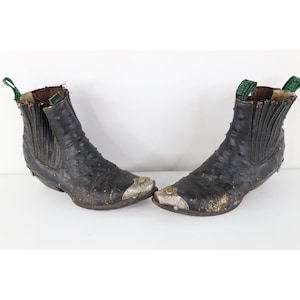 May include: A pair of black leather ankle boots with a textured finish, featuring silver-toned metal toe caps with engraved detailing, elastic side panels, and green pull tabs at the top. The boots show signs of wear on the leather surface.