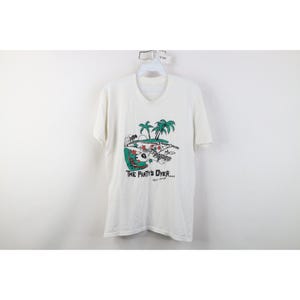 以下が含まれることがあります： Vネックの白い半袖Tシャツ。前面には、ヤシの木、サッカーボール、そして「THE PARTY'S OVER...」というテキストが描かれたトロピカルなシーンのグラフィックが描かれています。