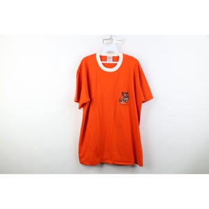 May include: A bright orange short-sleeve t-shirt with a white crew neck collar. The shirt features a small graphic on the lower right side depicting a black and white illustration of a cat with the text "Night Creatures" printed below it.