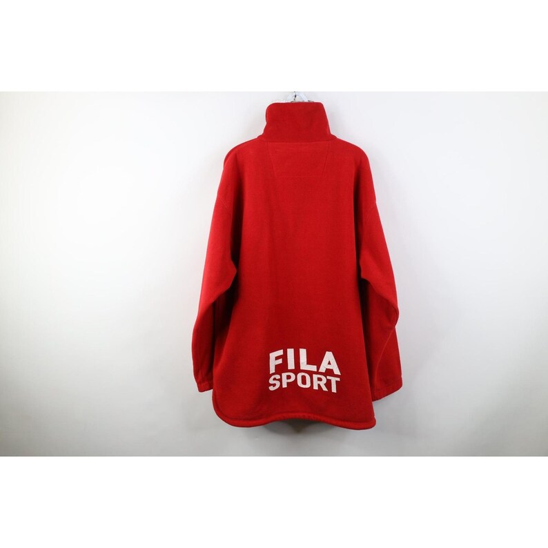 May include: A red fleece pullover with a high collar. The back of the garment features the white text "FILA SPORT". The long-sleeved top is hanging against a white background.