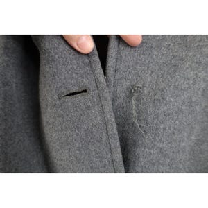 May include: Close-up of a gray wool coat with a button and a small hole with loose threads. The coat's fabric is a solid, medium gray color.