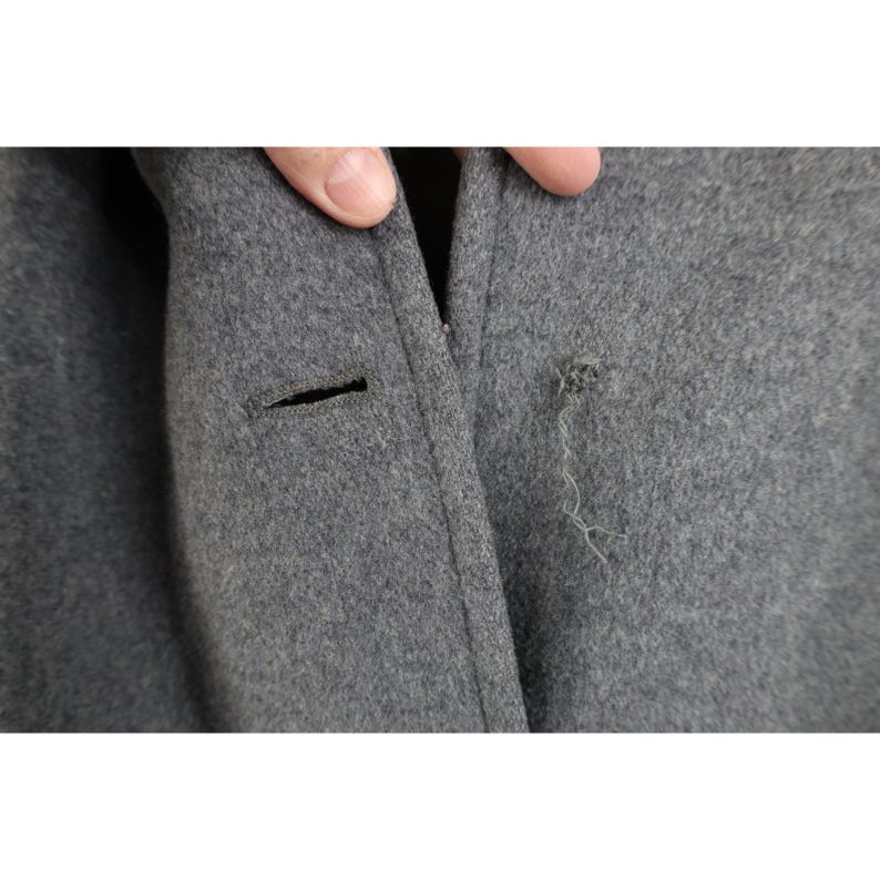 May include: Close-up of a gray wool coat with a button and a small hole with loose threads. The coat's fabric is a solid, medium gray color.
