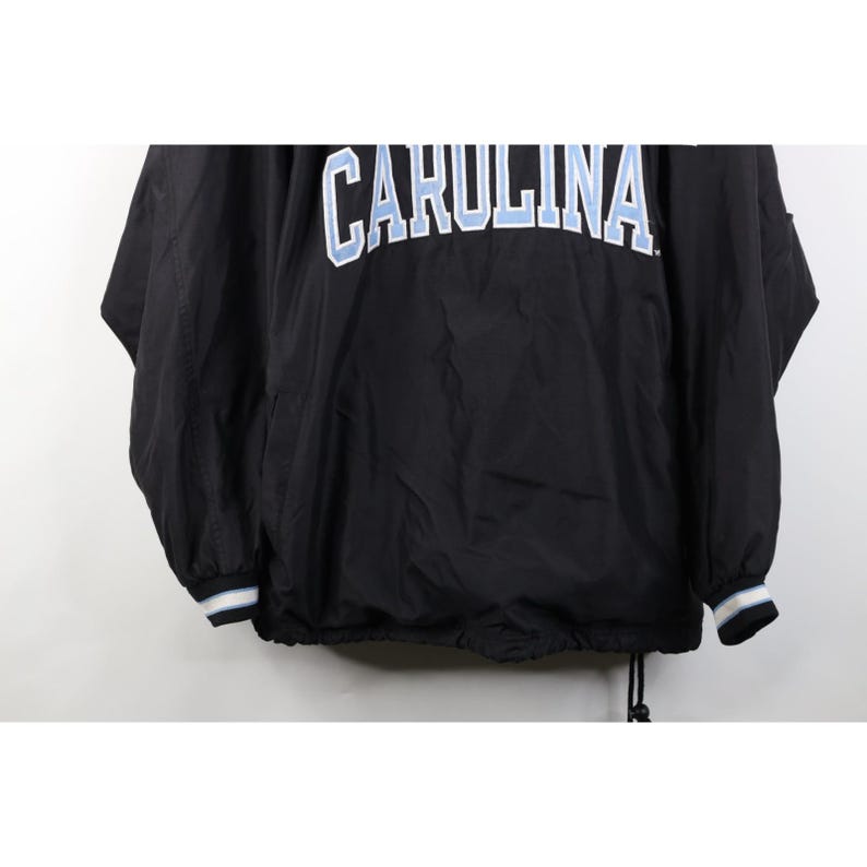 May include: Black jacket with the word "CAROLINA" in white and light blue lettering across the chest. The jacket has a drawstring at the bottom and white and blue striped cuffs.