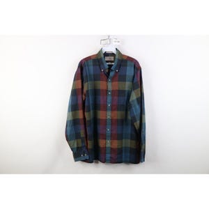 Puede incluir: Una camisa abotonada de manga larga con un estampado de cuadros. La camisa presenta una paleta de colores de azul, verde, rojo y marrón. La camisa está colgada de una percha blanca sobre un fondo blanco.