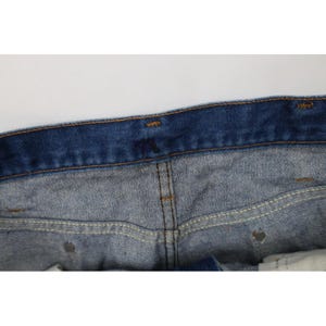 Op de afbeelding: Close-up van de tailleband en het bovenste deel van een blauwe spijkerbroek. De jeans heeft een klassiek ontwerp met vijf zakken, een knoopsluiting en een bruine gestikte naad. De denim heeft een vervaagde uitstraling.