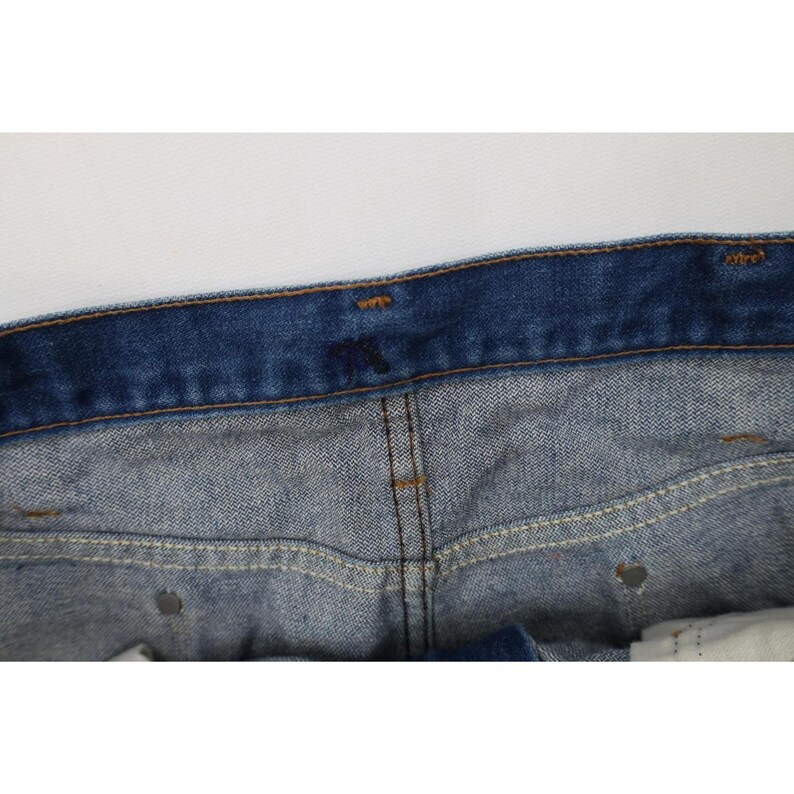 Op de afbeelding: Close-up van de tailleband en het bovenste deel van een blauwe spijkerbroek. De jeans heeft een klassiek ontwerp met vijf zakken, een knoopsluiting en een bruine gestikte naad. De denim heeft een vervaagde uitstraling.