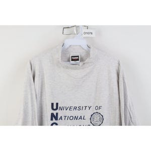 Op de afbeelding: Een lichtgrijs shirt met lange mouwen en een opstaande kraag. Het shirt toont de tekst "University of National" in marineblauw, samen met een cirkelvormig embleem. Het shirt hangt aan een witte hanger.