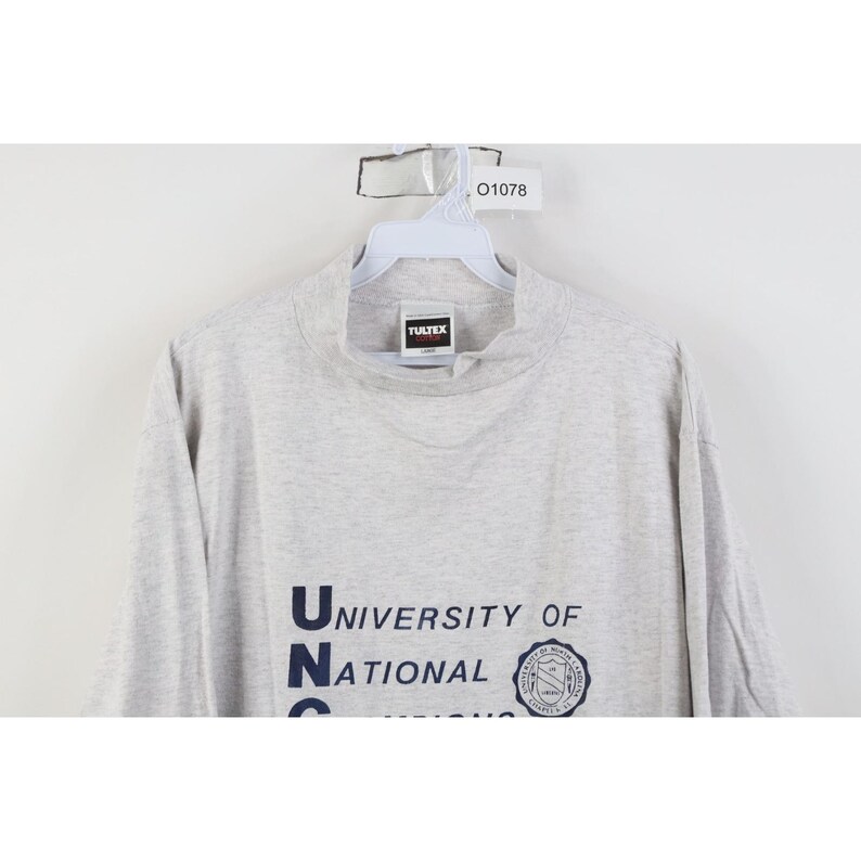 Op de afbeelding: Een lichtgrijs shirt met lange mouwen en een opstaande kraag. Het shirt toont de tekst "University of National" in marineblauw, samen met een cirkelvormig embleem. Het shirt hangt aan een witte hanger.