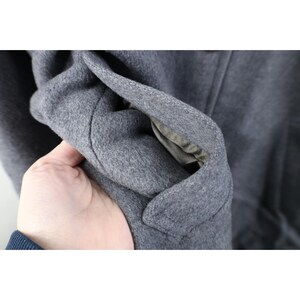May include: Close-up of a gray wool coat. The coat's fabric is a solid gray color, and the interior lining is visible. The coat's texture appears soft and warm, suggesting it's suitable for cold weather.