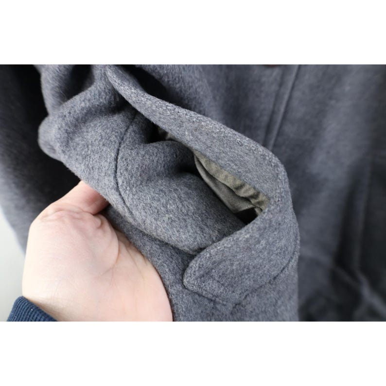 May include: Close-up of a gray wool coat. The coat's fabric is a solid gray color, and the interior lining is visible. The coat's texture appears soft and warm, suggesting it's suitable for cold weather.