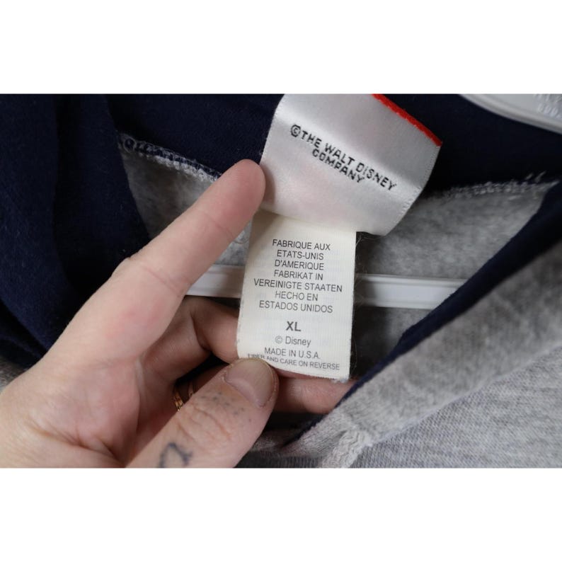 May include: A white clothing tag attached to a navy blue and heather gray garment. The tag features the text THE WALT DISNEY COMPANY, FABRIQUE AUX ETATS-UNIS D'AMERIQUE, FABRIKAT IN VEREINIGTE STAATEN, HECHO EN ESTADOS UNIDOS, size XL, and MADE IN U.S.A.