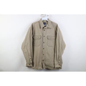May include: A beige button-down shirt with two chest pockets and a collar. The shirt has long sleeves with buttoned cuffs and a GAP logo tag. The shirt is made of a durable fabric, suitable for casual wear.