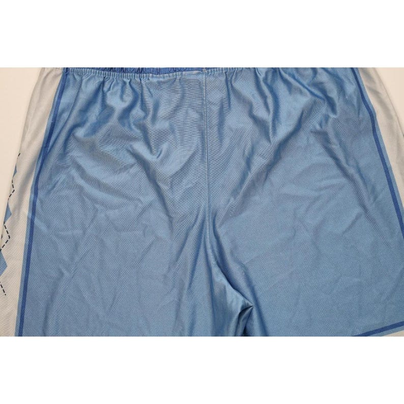 May include: Light blue athletic shorts featuring a smooth, synthetic fabric with a subtle mesh-like texture. The shorts include white side panels with a dark blue diamond pattern trim along the edges, an elastic waistband, and visible stitching.