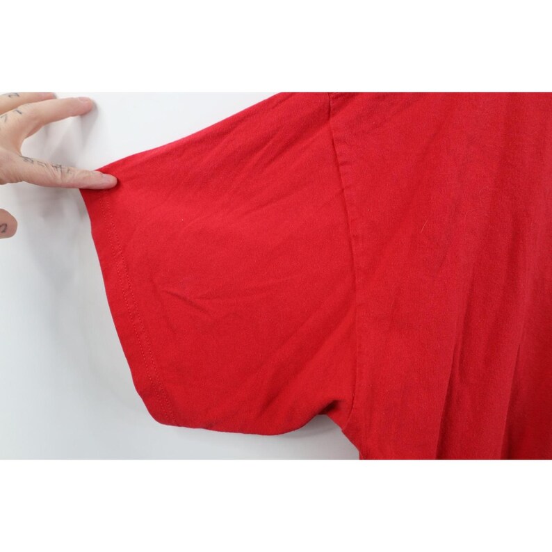 May include: Close-up of a bright red t-shirt sleeve. The fabric appears to be a soft cotton material. The sleeve is being held by a hand with tattoos.