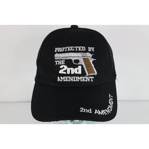 Peut inclure: Une casquette de baseball noire avec une broderie blanche indiquant PROTECTED BY THE 2nd AMENDMENT. Un motif brodé représentant une arme de poing argentée et marron est centré au-dessus du texte. 2nd AMENDMENT est brodé en blanc sur la visière.
