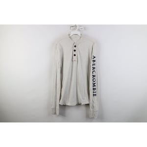 May include: A light grey long-sleeve henley shirt featuring a three-button placket at the neckline and the word ABERCROMBIE printed in dark navy blue capital letters vertically down the length of the left sleeve, hanging on a white hanger.