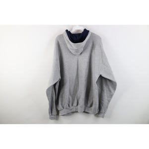 May include: A heather gray long-sleeve pullover hoodie featuring a contrasting dark navy blue interior lining within the hood. The garment is constructed with a standard drawstring-free hood, ribbed cuffs at the sleeves, and a ribbed hemline.