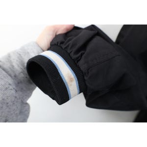 May include: Black jacket sleeve with a ribbed cuff. The cuff features a black base with white and light blue stripes. A brown stain is visible on the cuff. The jacket is held against a white background.