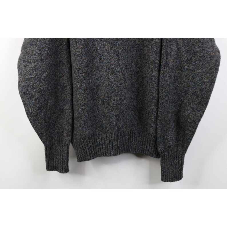 May include: A gray sweater with a speckled pattern. The sweater has a ribbed hem and cuffs. The fabric appears to be a blend of gray, brown, and blue yarns, creating a textured look. The sleeves are long and the overall style is casual.