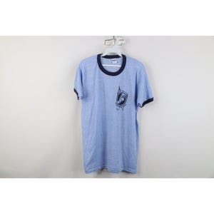 May include: Pale blue ringer t-shirt with navy trim on the collar and sleeves. The shirt has a graphic on the front with text and a design. The t-shirt is hanging on a white hanger.