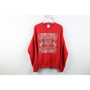 May include: A red crewneck sweatshirt with a graphic design. The design includes the text "Apples & Spice" and "American Slice" with a heart-shaped flag and apple illustrations. The sweatshirt is hanging on a white hanger.