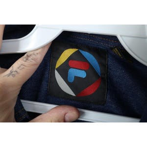 May include: Close-up of a dark blue denim jacket with a black label featuring the FILA logo. The logo is a stylized "F" in blue, red, and white, surrounded by a colorful geometric design. The jacket is hanging on a white hanger.