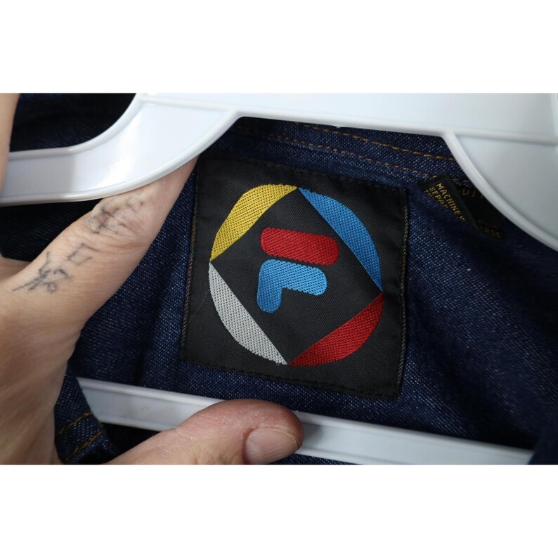 May include: Close-up of a dark blue denim jacket with a black label featuring the FILA logo. The logo is a stylized "F" in blue, red, and white, surrounded by a colorful geometric design. The jacket is hanging on a white hanger.