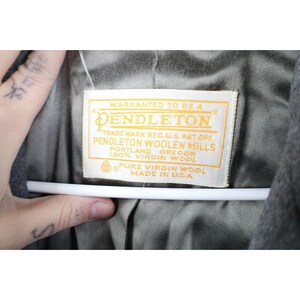 May include: Close-up of a Pendleton label, featuring text in a gold rectangular frame. The label reads "Warranted to be a Pendleton" and "Pendleton Woolen Mills, Portland, Oregon, 100% Virgin Wool, Made in U.S.A."
