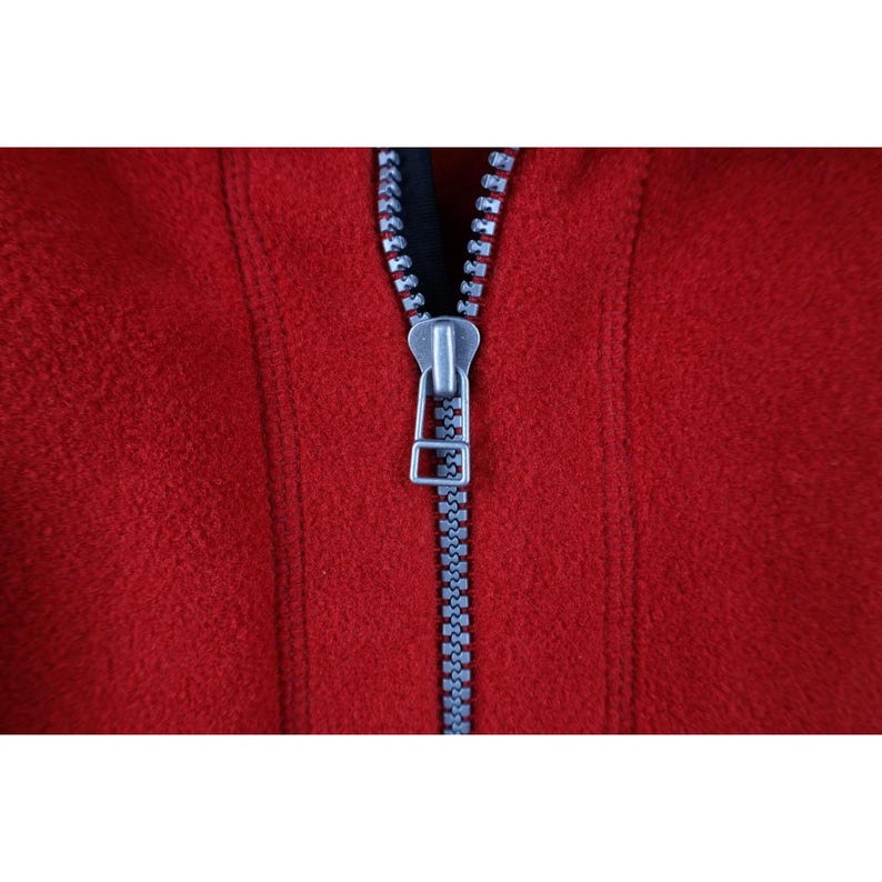 May include: Close-up of a red fleece jacket with a silver zipper. The zipper is centered and fully closed, with a black fabric visible behind it. The fleece has a soft, textured appearance.