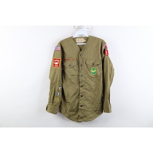 May include: An olive green long-sleeve button-down shirt featuring various embroidered patches on the sleeves and chest. The patches include a United States flag, a red lamp emblem, circular badges, and a green circular patch with a yellow symbol.
