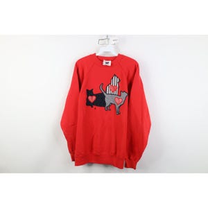 May include: Red sweatshirt with a cat design. The design features three cats in black, gray, and striped patterns, each with a heart. The sweatshirt has long sleeves and a crew neck.