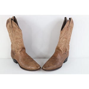 Puede incluir: Un par de botas vaqueras de cuero color canela con punta afilada, tacón apilado y patrones de costuras decorativas en la caña alta. Las botas tienen un acabado desgastado y un diseño clásico de calce fácil con tiradores laterales.