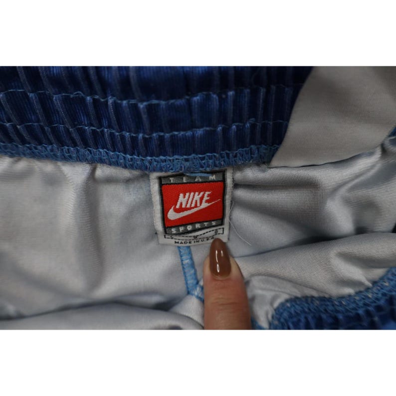 May include: Blue athletic shorts with an elastic waistband and a white inner lining. A rectangular Nike Team Sports tag is sewn onto the interior, featuring the Nike logo, the text TEAM SPORTS, and the words MADE IN U.S.A. at the bottom.