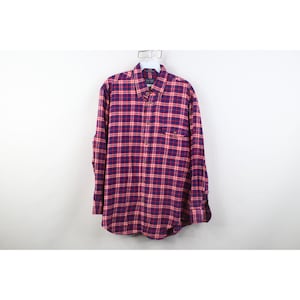 Puede incluir: Camisa abotonada de manga larga con estampado de cuadros rojos y morados. La camisa tiene un bolsillo en el pecho en el lado izquierdo y cuelga sobre un fondo blanco. El cuello está abotonado y las mangas están abrochadas.