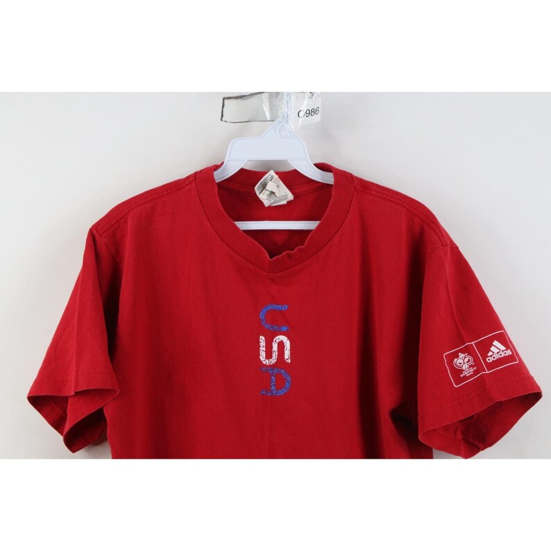 May include: Red t-shirt with the word "USA" in blue and white lettering. The Adidas logo is on the right sleeve. The shirt is hanging on a white hanger.