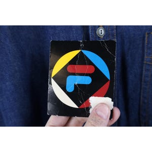 May include: Close-up of a dark blue denim shirt with a tag featuring the Fila logo. The tag is black with a colorful geometric design in yellow, red, blue, and white. The shirt has a button closure and a pocket.