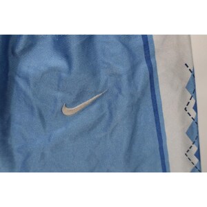 May include: Light blue athletic shorts featuring a silver Nike swoosh logo on the left side. The fabric has a smooth, slightly shiny texture. A white vertical side panel displays a blue zigzag pattern outlined with thin black dashed lines.