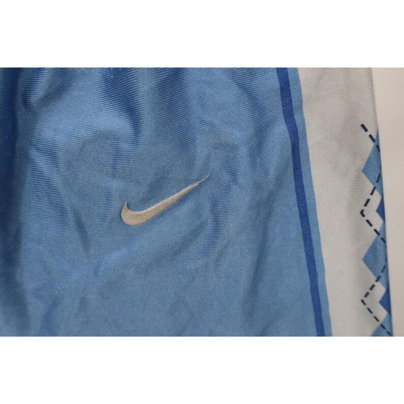 May include: Light blue athletic shorts featuring a silver Nike swoosh logo on the left side. The fabric has a smooth, slightly shiny texture. A white vertical side panel displays a blue zigzag pattern outlined with thin black dashed lines.
