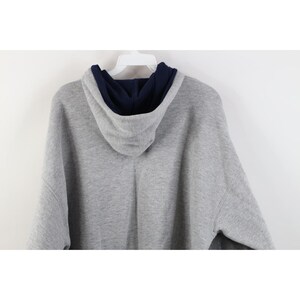 May include: A heather gray long-sleeve hooded sweatshirt featuring a contrasting dark navy blue interior lining within the hood. The garment is displayed on a white plastic hanger, showing the back view of the hood and the upper shoulder area.
