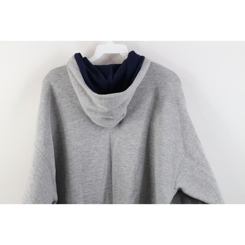 May include: A heather gray long-sleeve hooded sweatshirt featuring a contrasting dark navy blue interior lining within the hood. The garment is displayed on a white plastic hanger, showing the back view of the hood and the upper shoulder area.