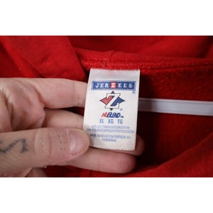Peut inclure: Gros plan d'un sweat &agrave; capuche rouge avec une &eacute;tiquette de la marque Jerzees. L'&eacute;tiquette affiche le logo de la marque, les informations de taille (XL, XG, TG) et la composition du tissu : 50% coton et 50% polyester. L'&eacute;tiquette indique &eacute;galement que l'article a &eacute;t&eacute; fabriqu&eacute; au Honduras.