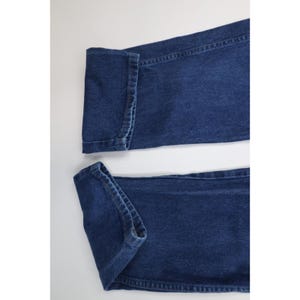 May include: Close-up of two sections of blue denim jeans. The jeans are folded at the bottom hem, showing the inside stitching and the texture of the denim fabric. The background is plain white.