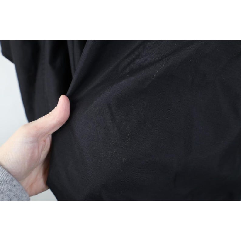 May include: Close-up of black fabric with visible texture and slight discoloration. The fabric is being held, revealing its folds and the way it drapes. The image highlights the material's dark color and surface details.