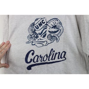 Op de afbeelding: Een lichtgrijs sweatshirt met een marineblauwe afbeelding van een rammenkop met de letters "UNC" op een hoed. Onder de afbeelding staat het woord "Carolina" in marineblauwe letters.